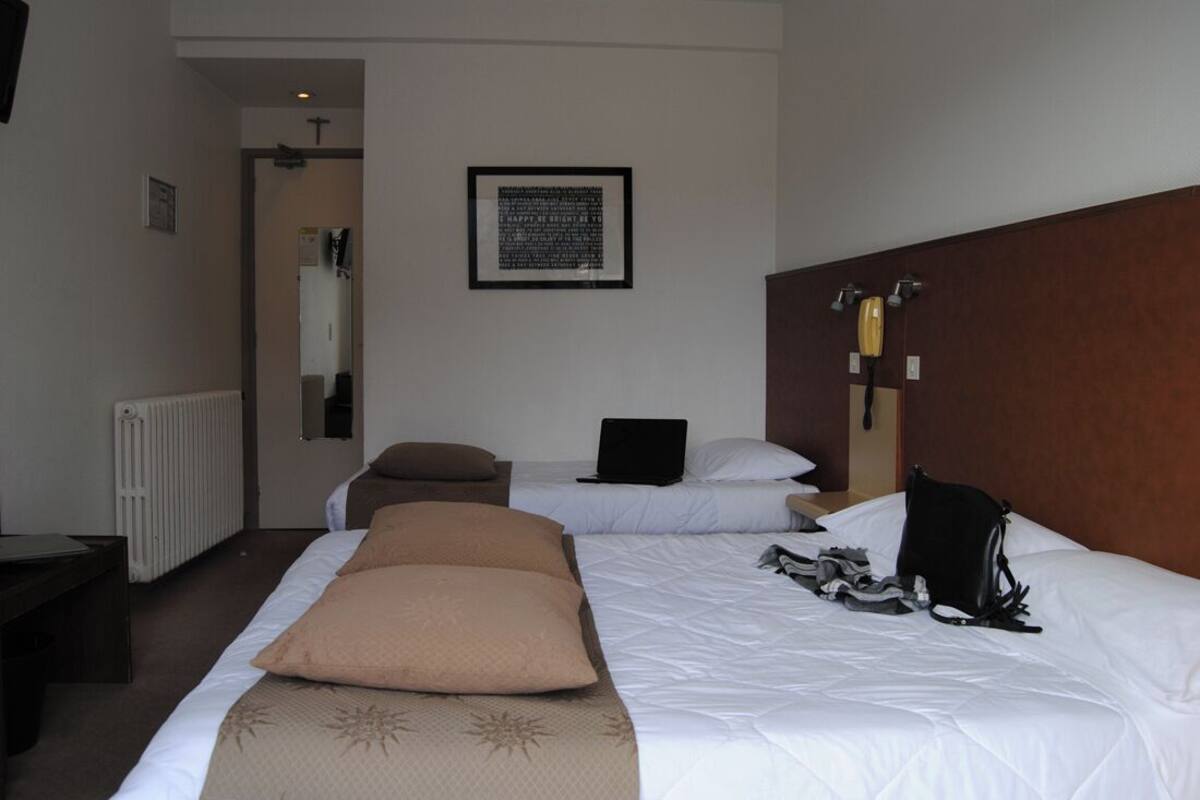 A hotel room with two beds, a desk, a chair, a radiator, a framed quote on the wall, and a telephone.