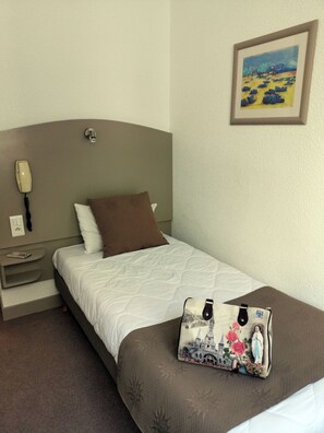 Habitación de hotel con cama, teléfono en la mesita de noche, lámpara de noche, almohada, un cuadro enmarco y un bolso decorativo.