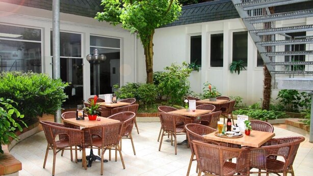 An outdoor patio area with wicker tables and chairs, surrounded by greenery and a staircase. - Adonis Lourdes Notre Dame De La Sarte (Lourdes)