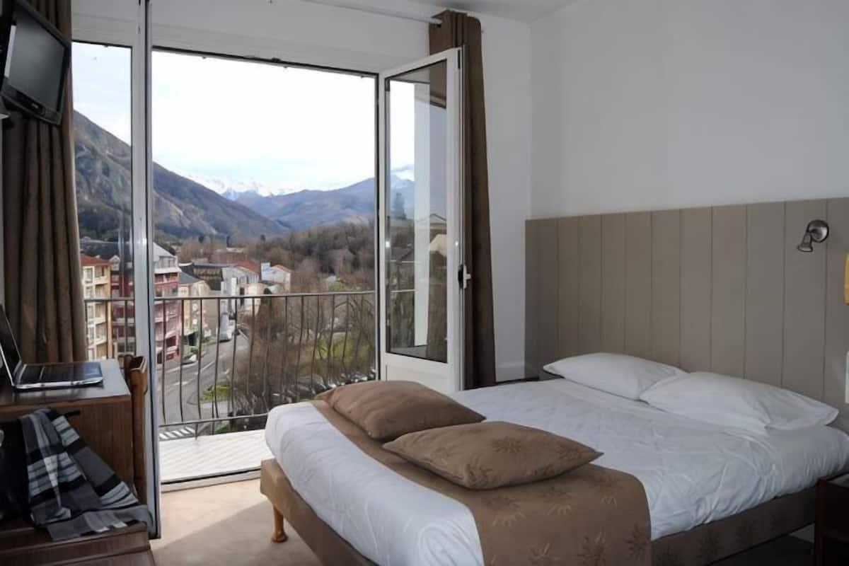 A bedroom with a bed, a laptop on a desk, and a view of mountains through a balcony.