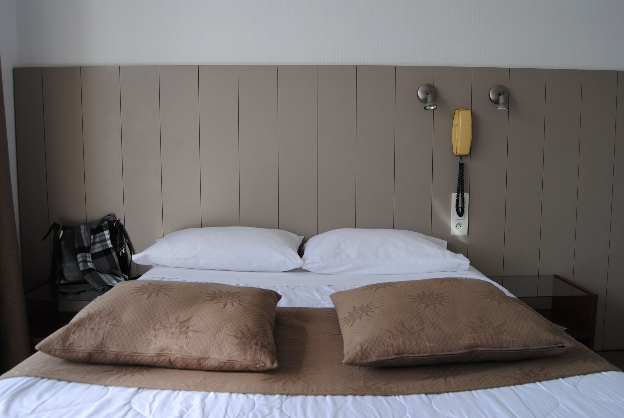 A hotel room with a bed, two pillows, a blanket, a telephone, and a wall-mounted lamp.