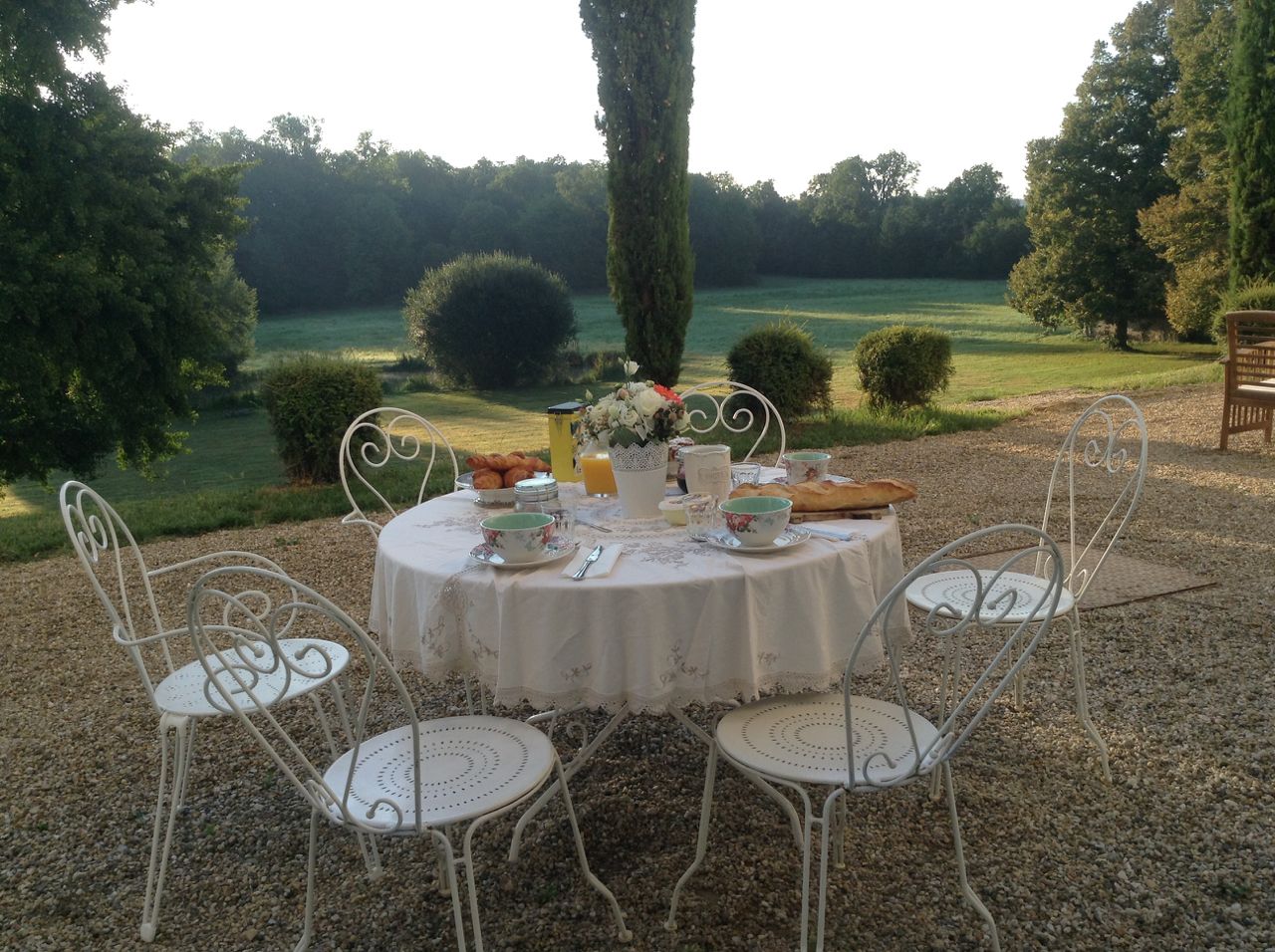 Une table ronde installée à l’extérieur, entourée de chaises en métal blanc, et le tout posé sur du gravier.