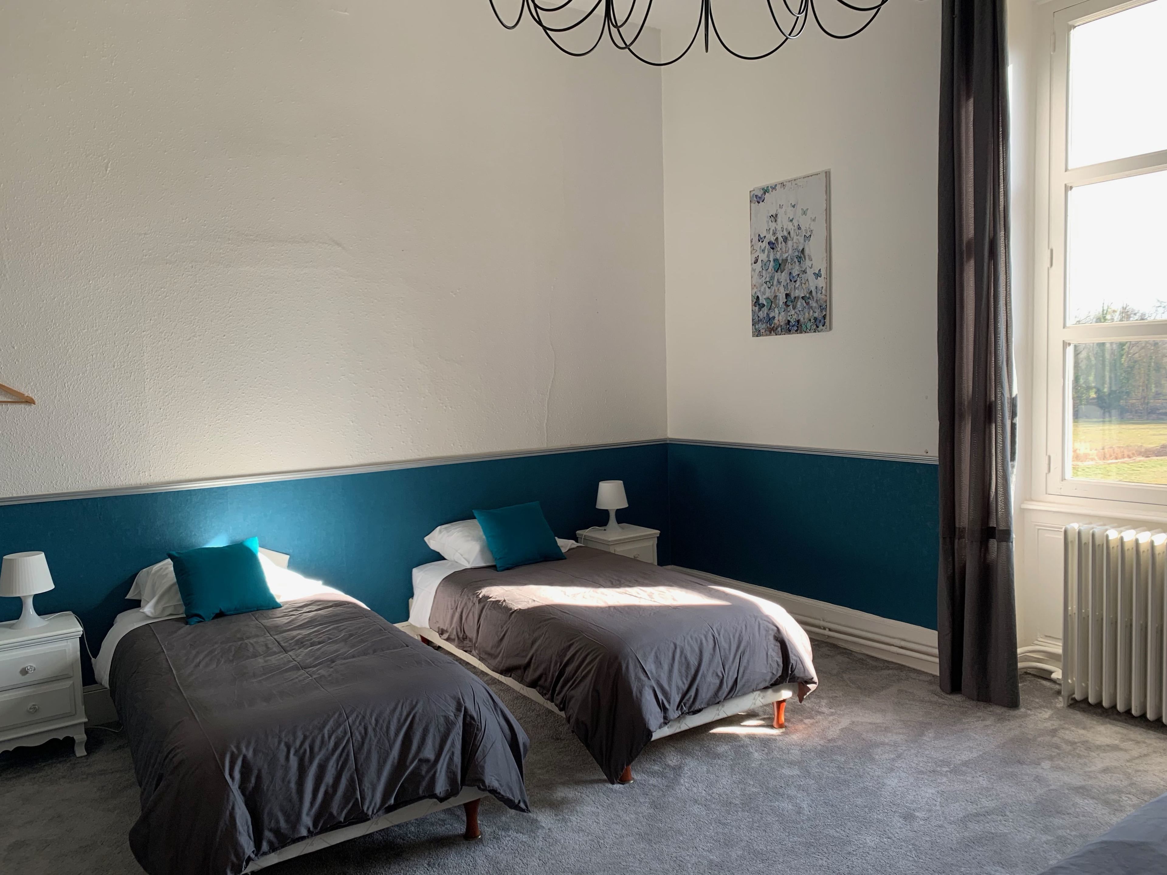Een slaapkamer met twee bedden, een nachtkastje met een lamp, een schilderij aan de muur en een raam met gordijnen.