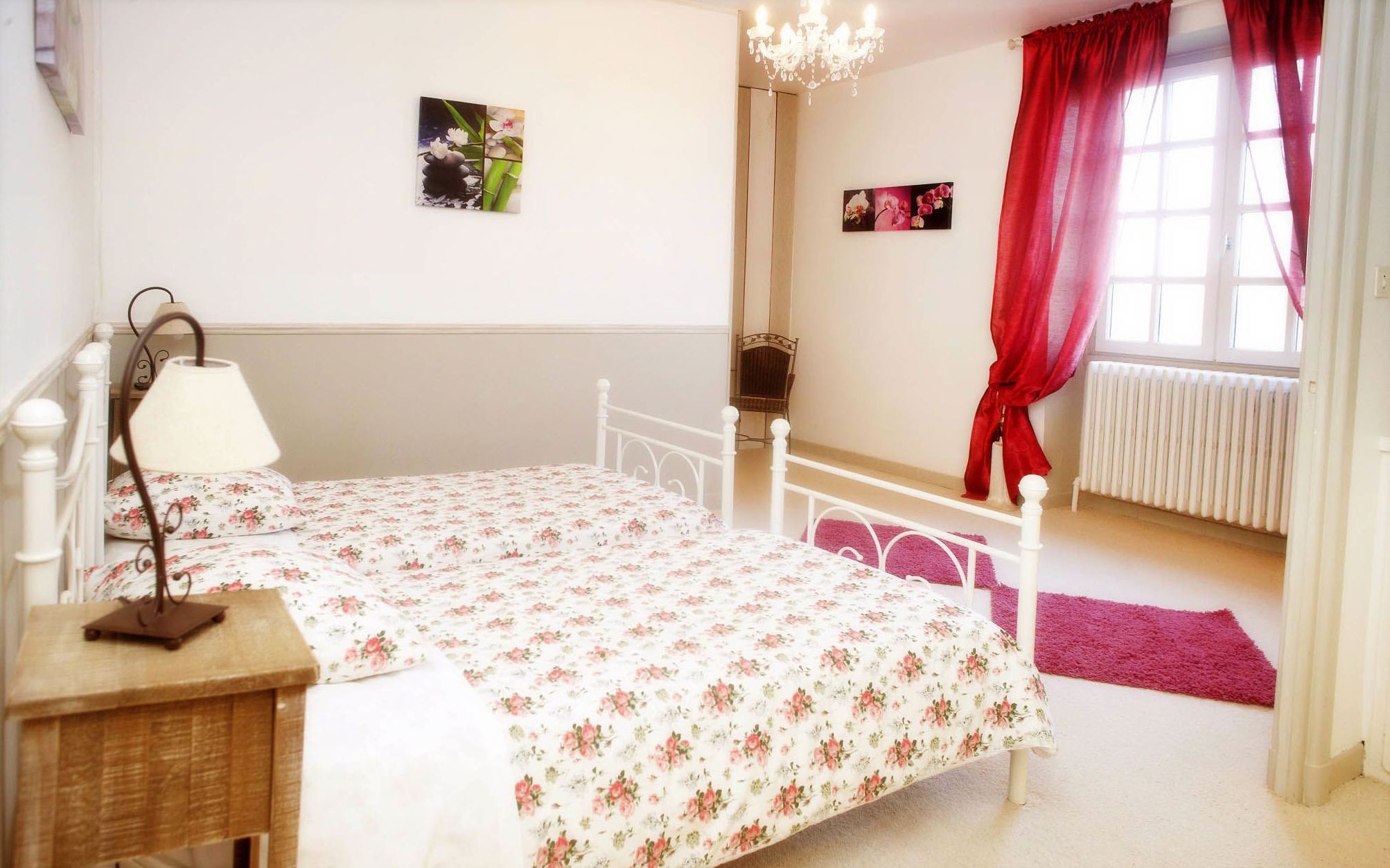 A bedroom with a bed, a nightstand, a chair, a radiator, and a window with red curtains.