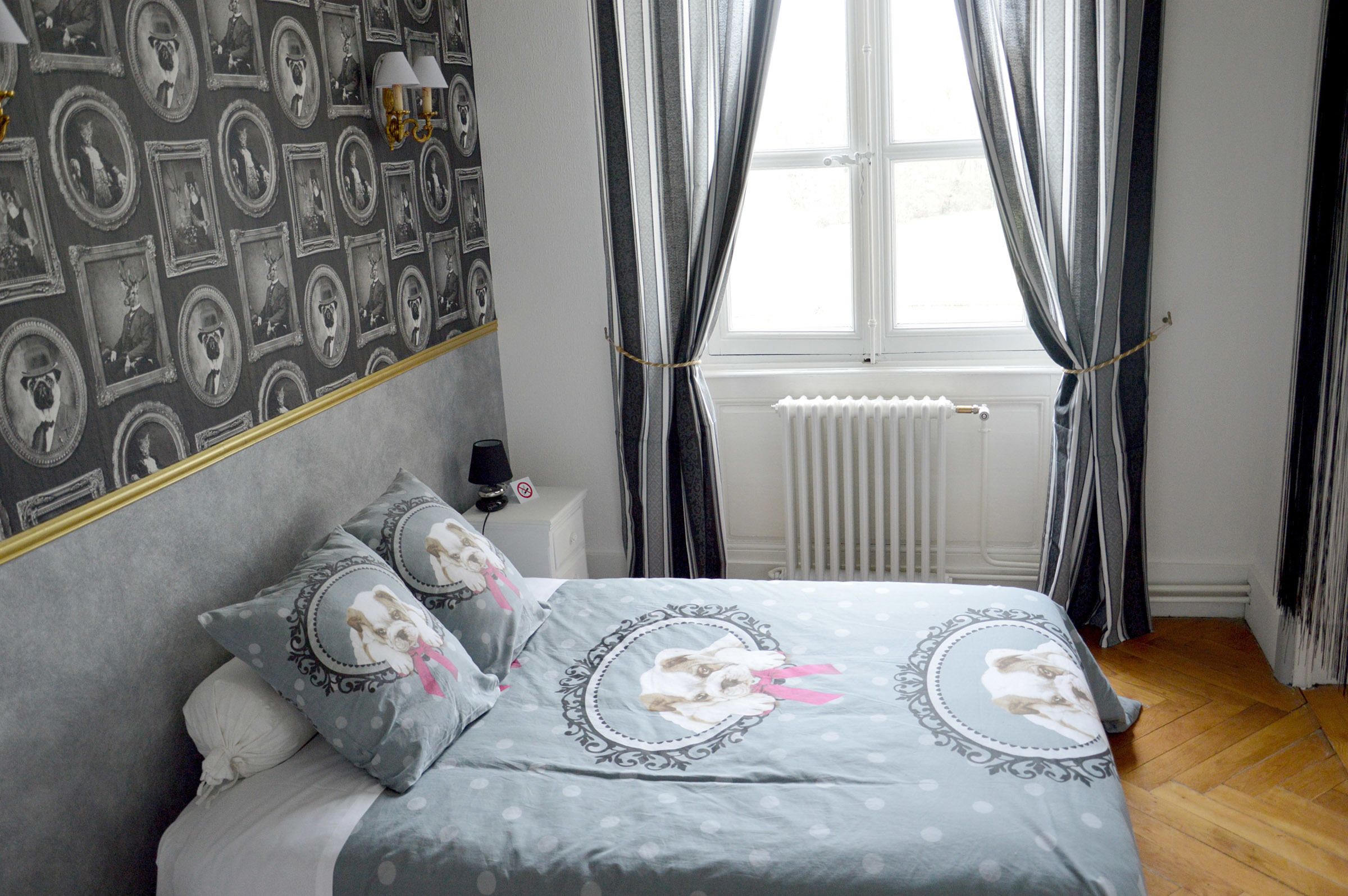 Un dormitorio con cama, almohadas, mesita de noche, lámpara, ventana con cortinas y radiador.