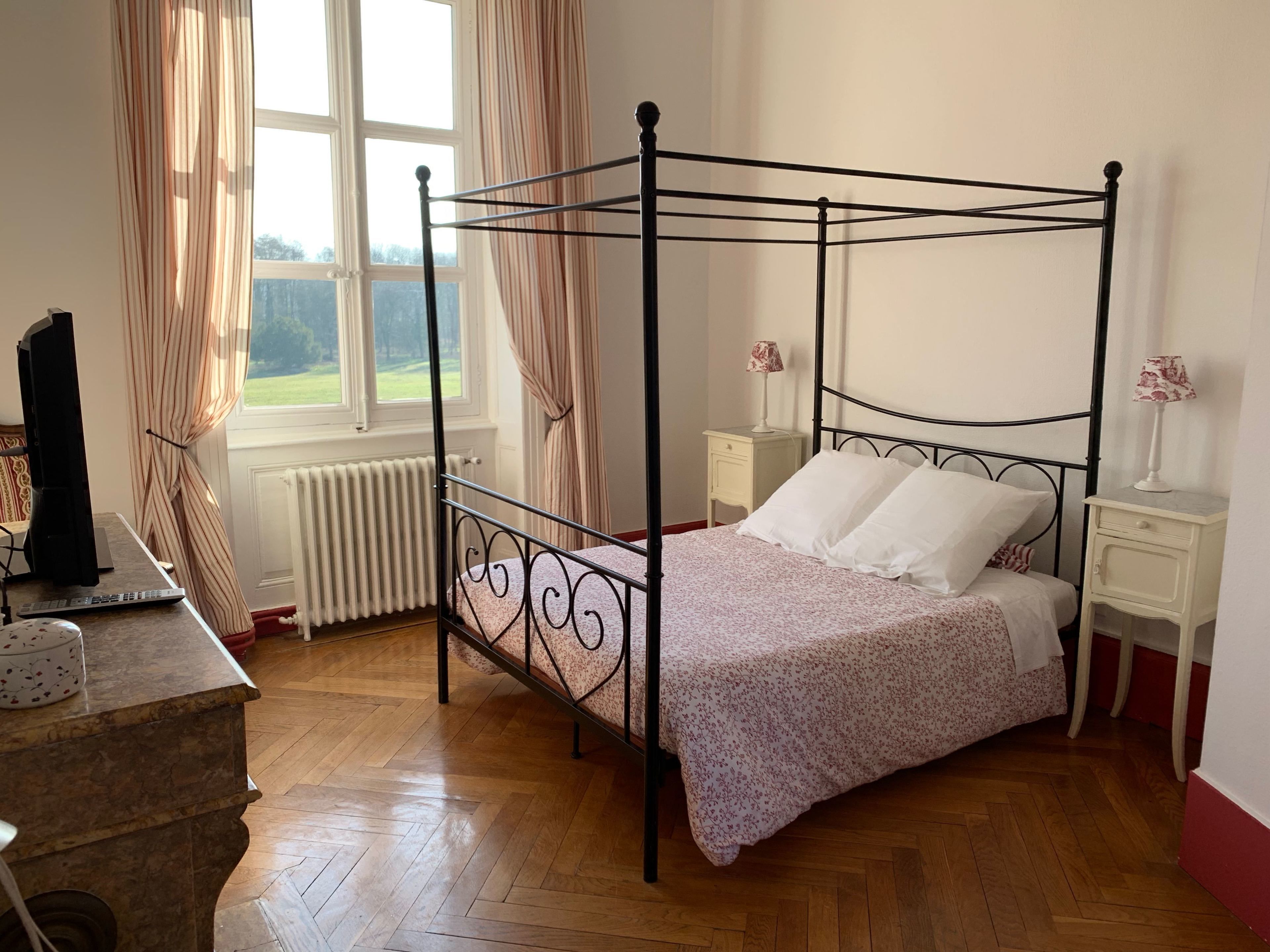 A four-poster bed with a floral bedspread, two bedside tables with lamps, a radiator, and a window with curtains.