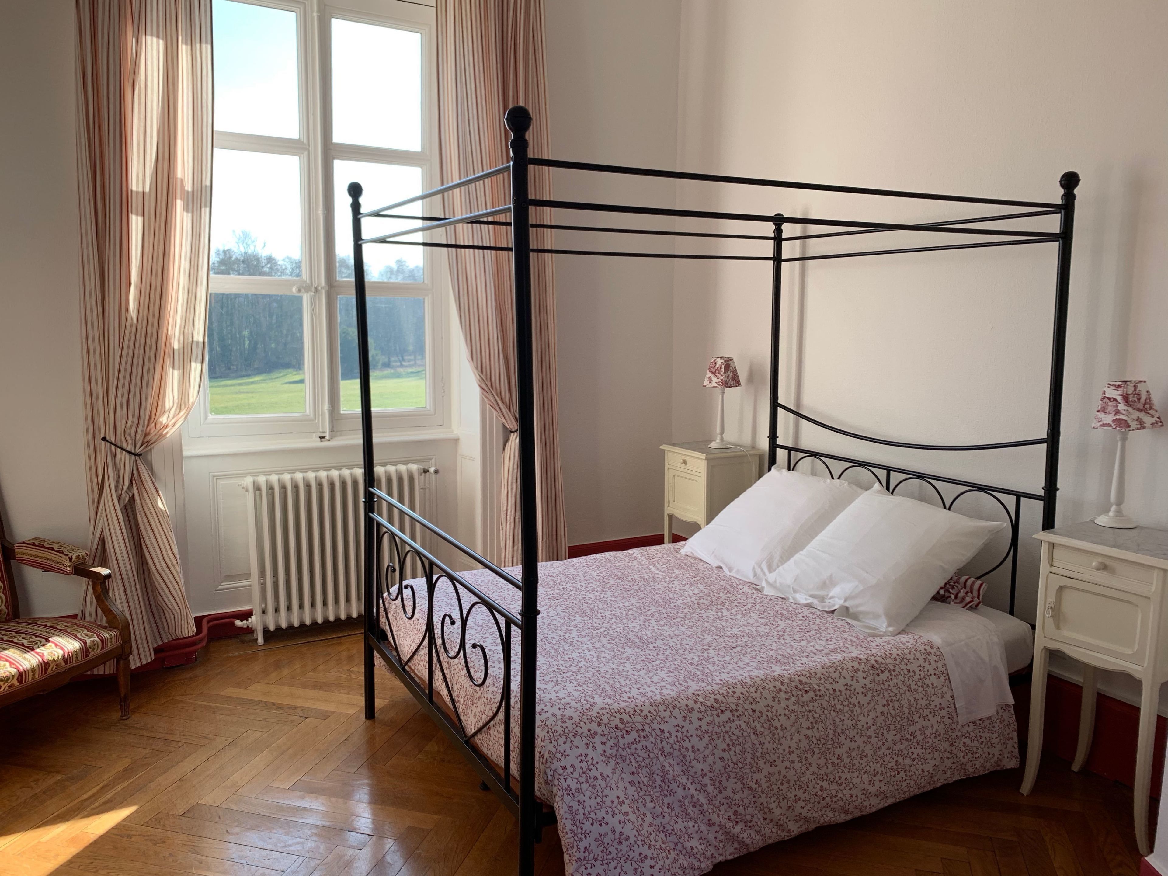 Een hemelbed met een bloemenbedekking, een nachtkastje met een lamp, en uitzicht op een groen landschap door een raam.
