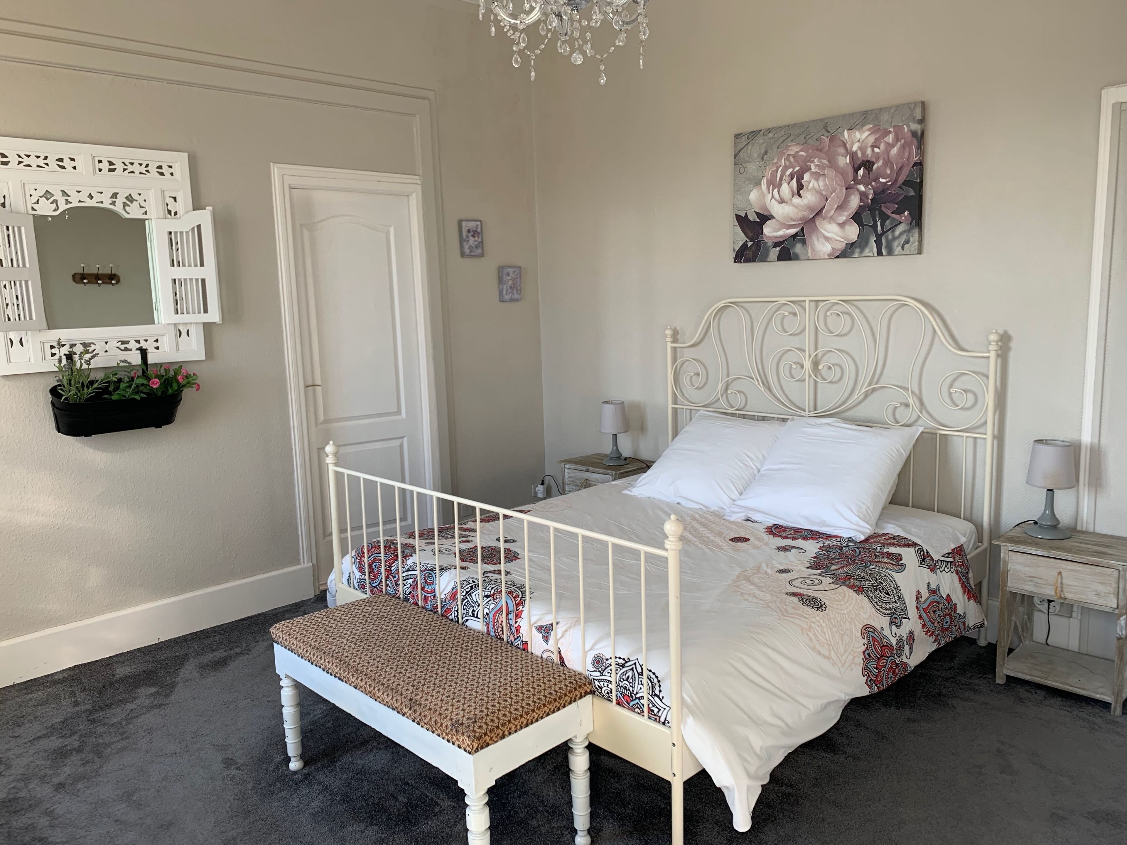 A bedroom with a white metal bed frame, a bench, a nightstand, a lamp, a wall-mounted mirror, and a floral painting.