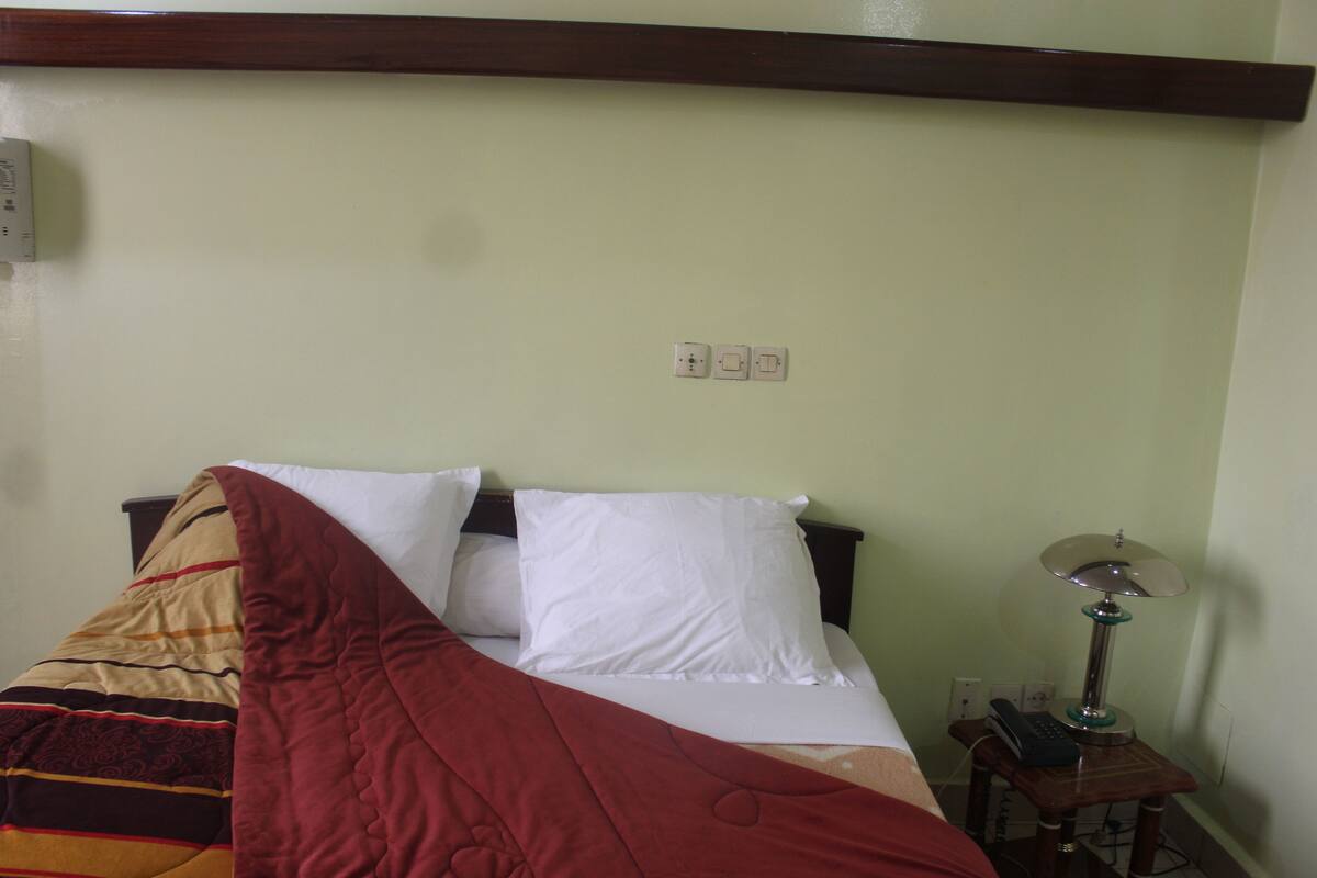 Una cama bien hecha con una manta roja, una lámpara de noche y un enchufe de pared.