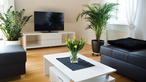 A modern living room with a flat-screen TV on a white stand, a grey sofa, a black ottoman, a white coffee table with a vase of yellow flowers, and two potted plants.