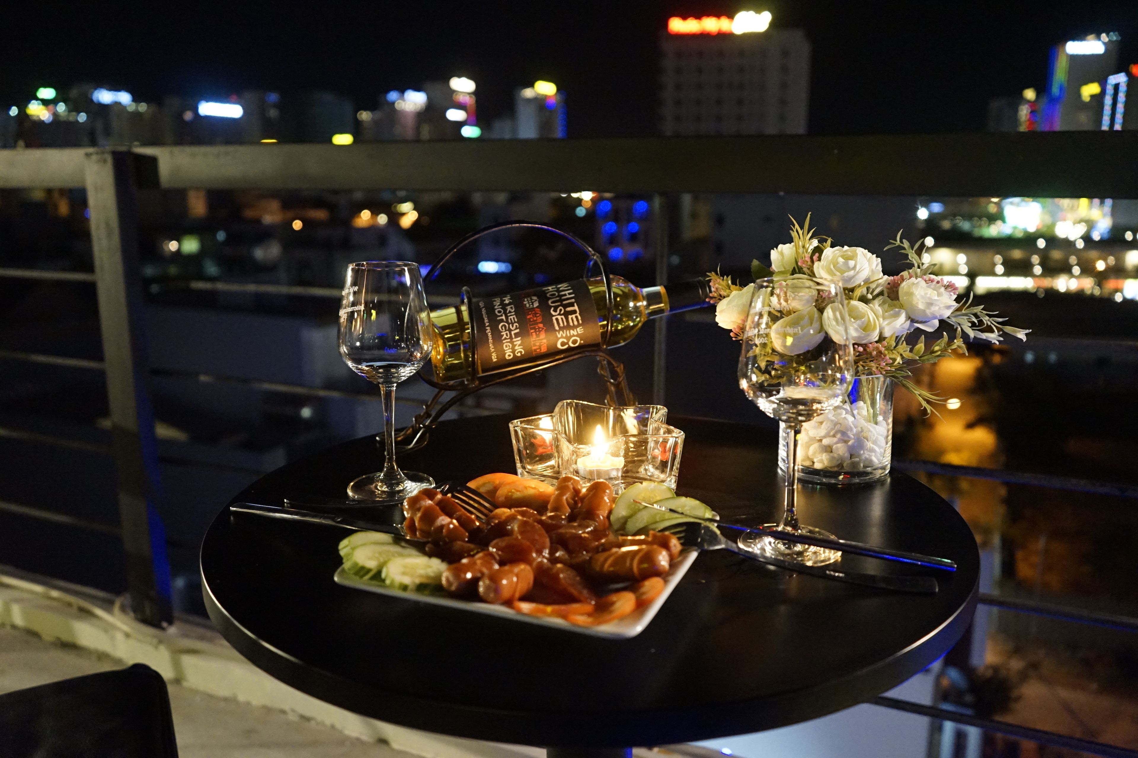 A table set with a bottle of white rum, a glass of water, a candle, and a plate of shrimp on a balcony overlooking a cityscape at night.