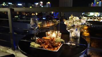 A table set with a bottle of white rum, a glass of water, a candle, and a plate of shrimp on a balcony overlooking a cityscape at night.
