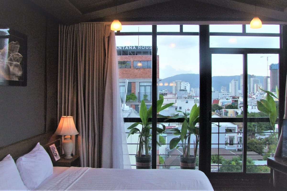 A hotel room with a bed, a nightstand with a lamp, a window with curtains, and a view of the cityscape.