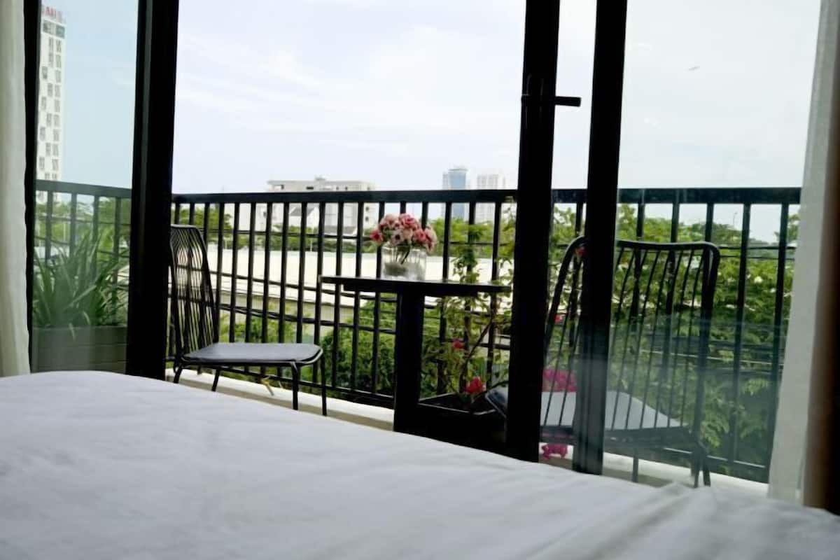 A hotel room with a bed, a small table with a vase of flowers, and a balcony with a view of buildings and greenery.