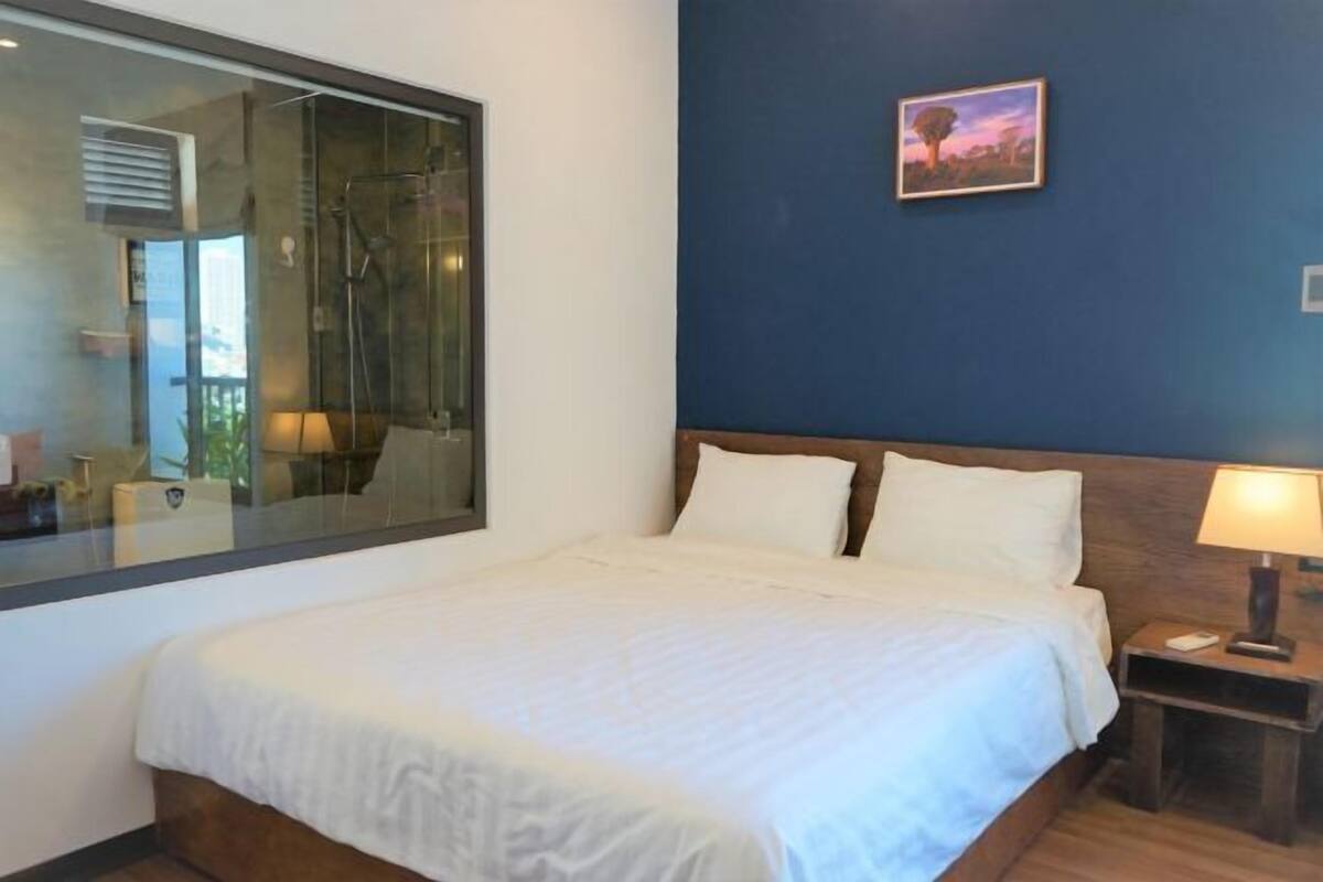 A bedroom with a large window, a bed with white linens, a nightstand with a lamp, and a painting on the wall.