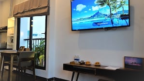 A hotel room with a large TV displaying a tropical beach scene, a desk with a tray of items, a chair, and a view of the outside balcony. - Oasis Hotel & Apartment (Da Nang)