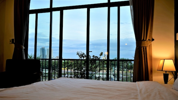 A hotel room with a large window, a bed, a nightstand, and a lamp. - Oasis Hotel & Apartment (Da Nang)