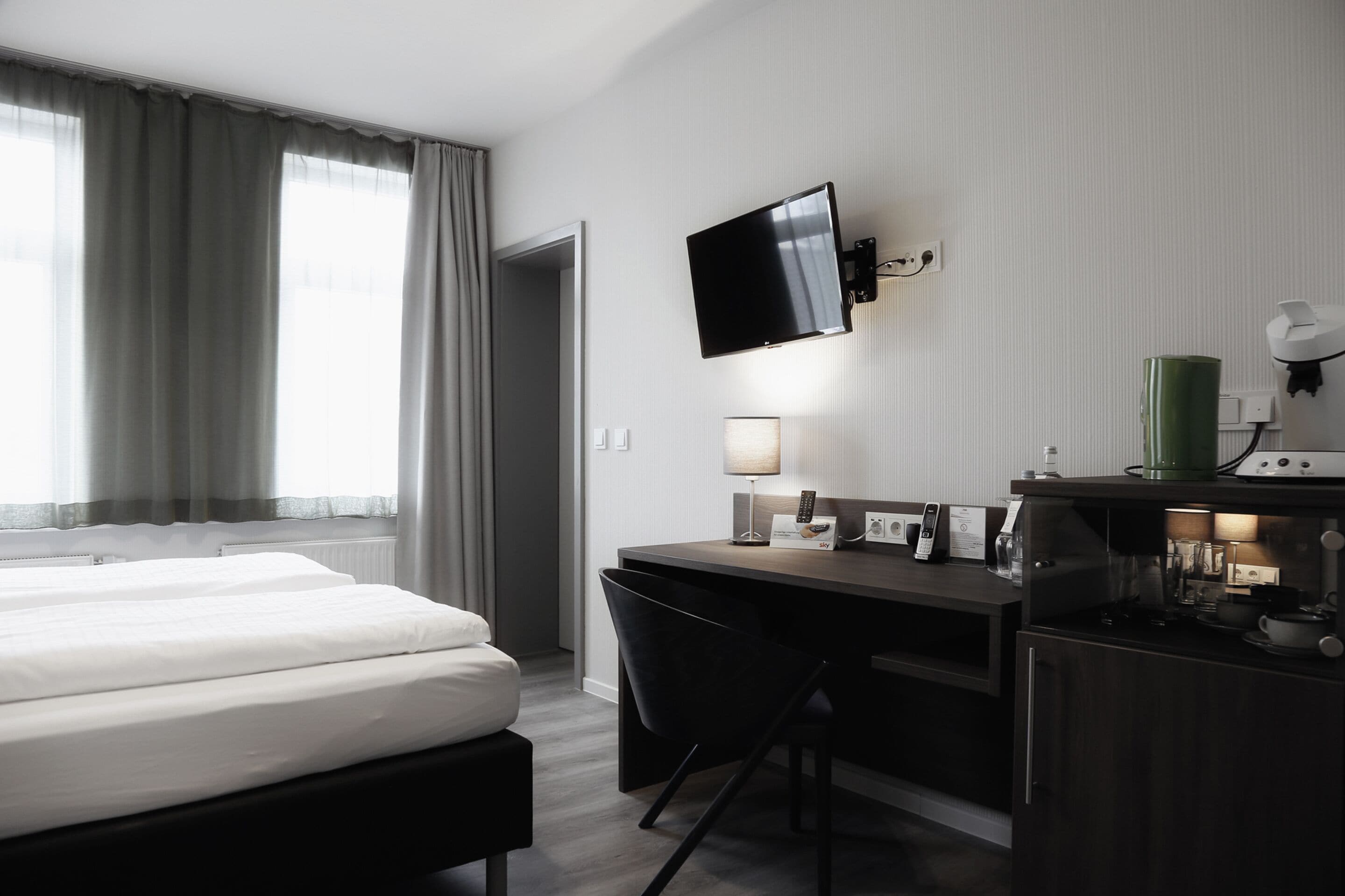 A hotel room with a bed, desk, chair, TV, lamp, and kitchenette.