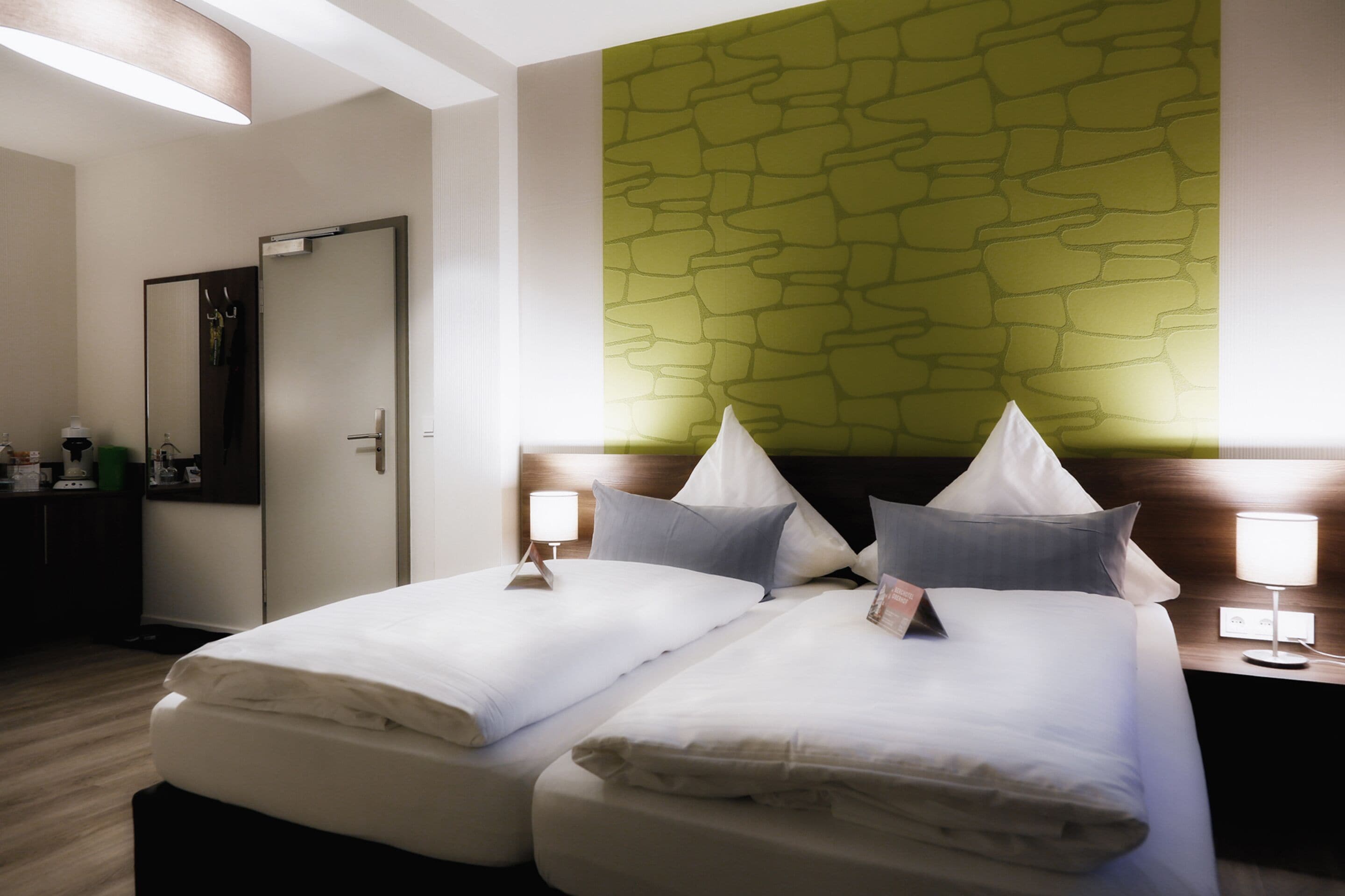 Een hotelkamer met twee bedden, een spiegel, een nachtkastje met een lamp, en een groene, structuurrijke muur.