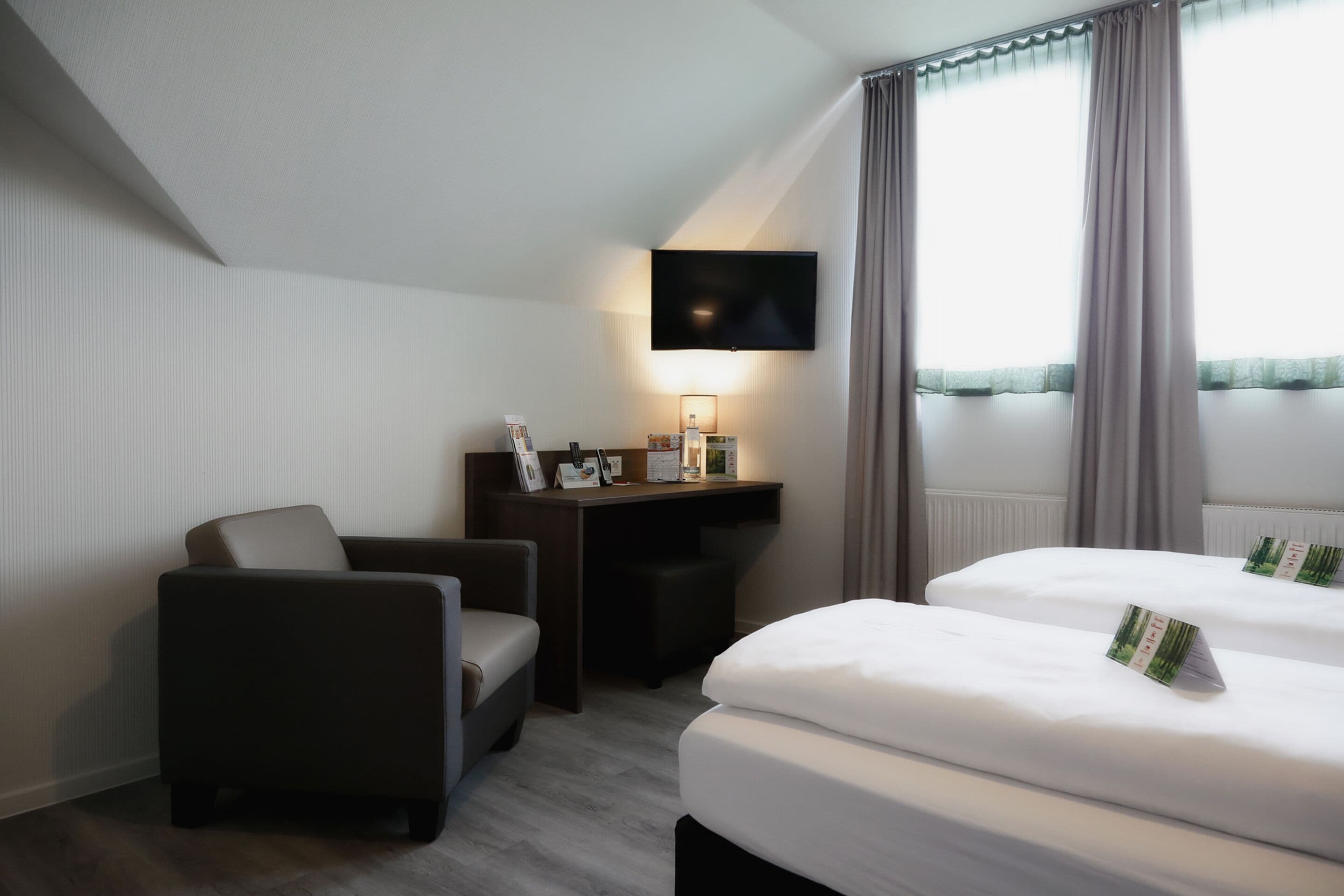 Habitación de hotel con cama, escritorio, silla, televisor y ventana con cortinas.
