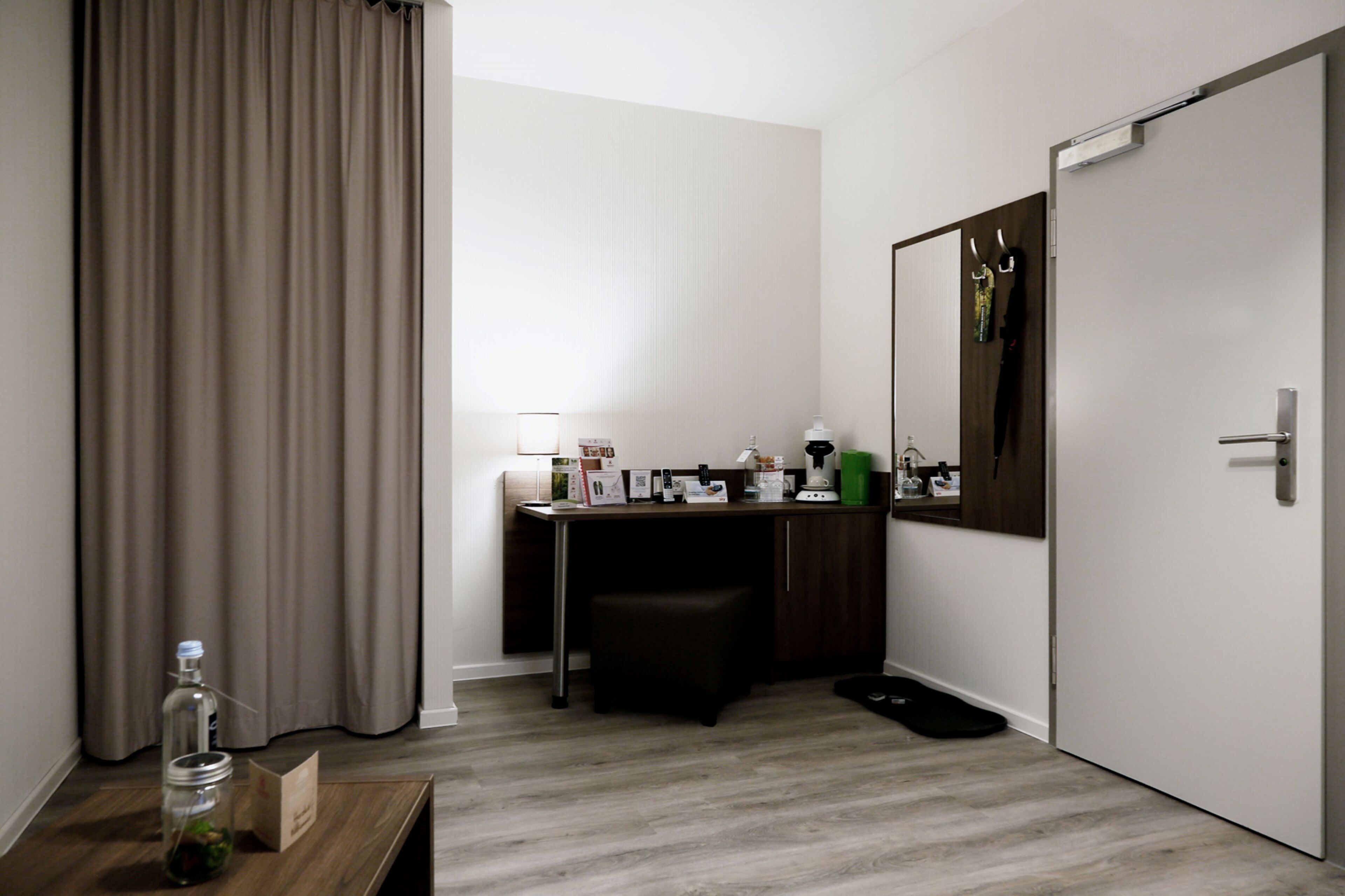 Une chambre d’hôtel équipée d’un bureau, d’un miroir, d’une porte, d’une fenêtre avec des rideaux et d’une petite table sur laquelle se trouvent une bouteille et une carte.