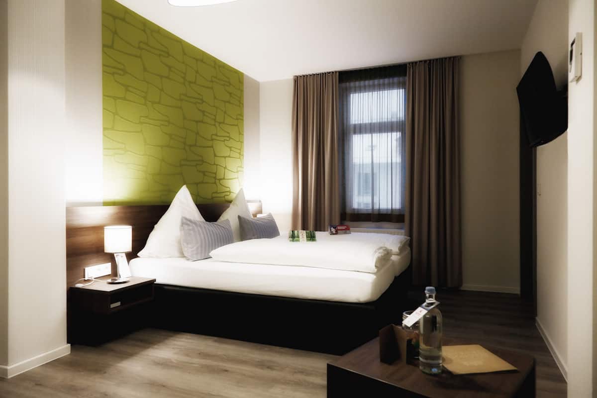 A hotel room with a bed, a nightstand, a lamp, a bottle, and a window with curtains.