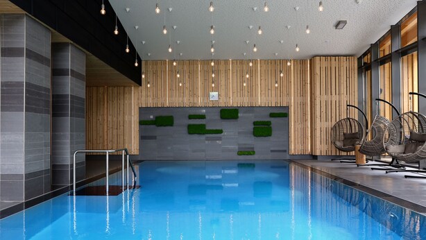 Ein modernes Hallenbad mit klarem, blauem Wasser, Holzvertäfelungen und Sitzbereichen mit Korbstühlen.