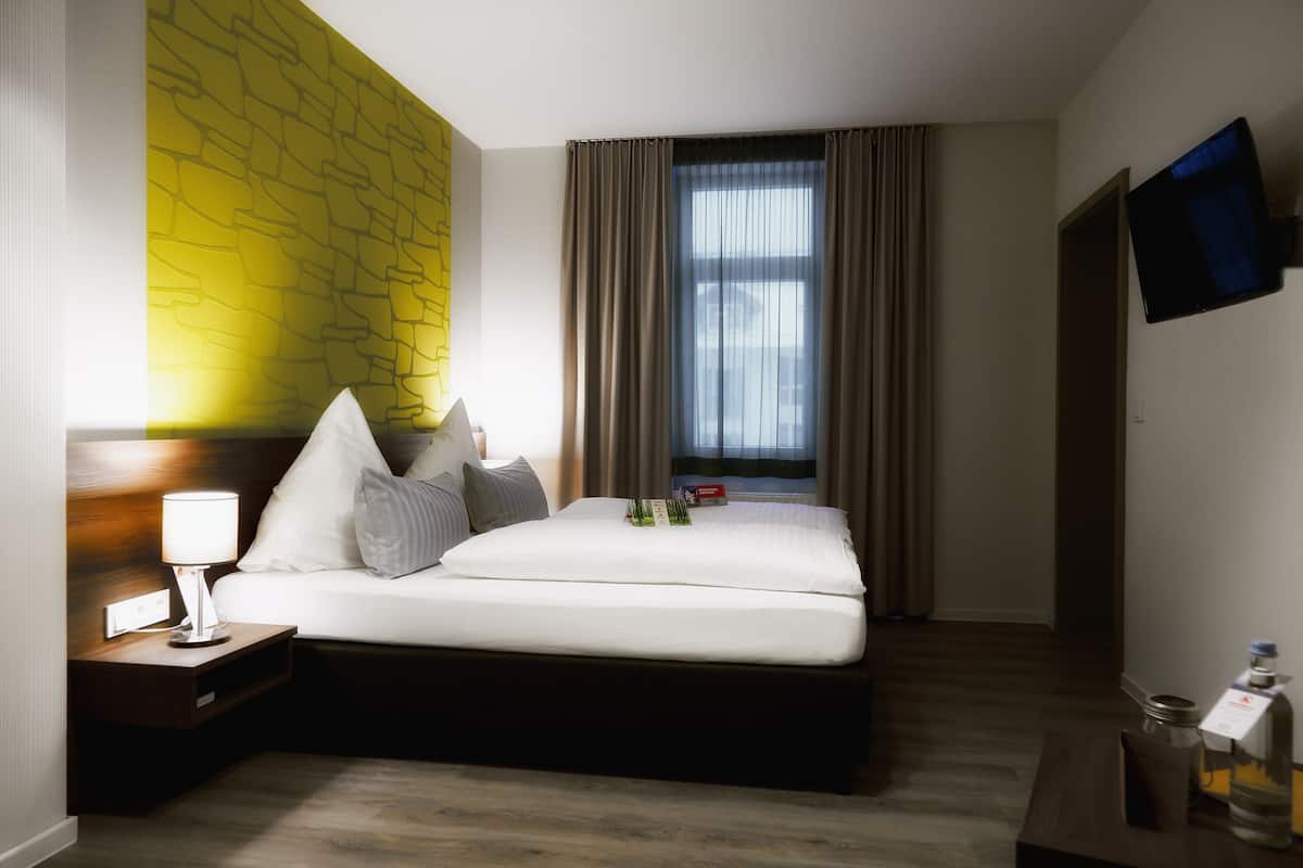 A hotel room with a bed, a nightstand with a lamp, a flat-screen TV mounted on the wall, and a window with curtains.