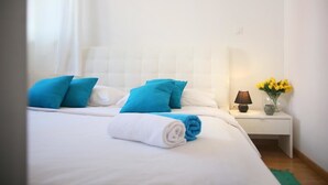 A neatly made bed with white and blue bedding, a rolled white towel, and a bedside table with a lamp and a vase of yellow flowers. - Apartment Marieta (Split)