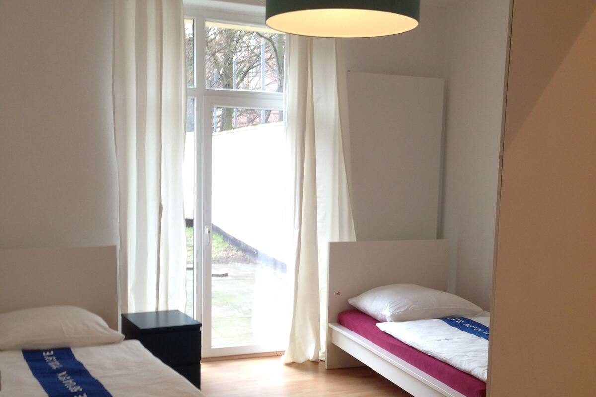 Un dormitorio con dos camas, una ventana con cortinas, piso de madera y una lámpara colgante.