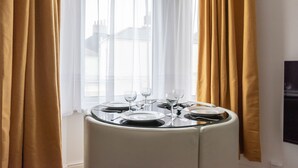Mesa de comedor para cuatro personas, con platos y vasos blancos, ubicada cerca de una ventana con cortinas blancas traslúcidas.