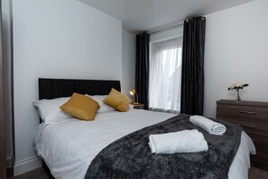 Una cama bien hecha con sábanas blancas y dos almohadas, un cabecero oscuro, una mesita de noche con una lámpara y un aparador con un jarrón de flores.