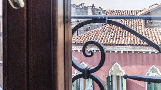 A view through a window with a decorative iron grille, revealing a building with terracotta roof tiles and green shutters. - Maurizio House (Venice)