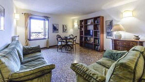 A living room with a sofa, a dining table with chairs, a bookshelf, and a window with curtains. - Maurizio House (Venice)