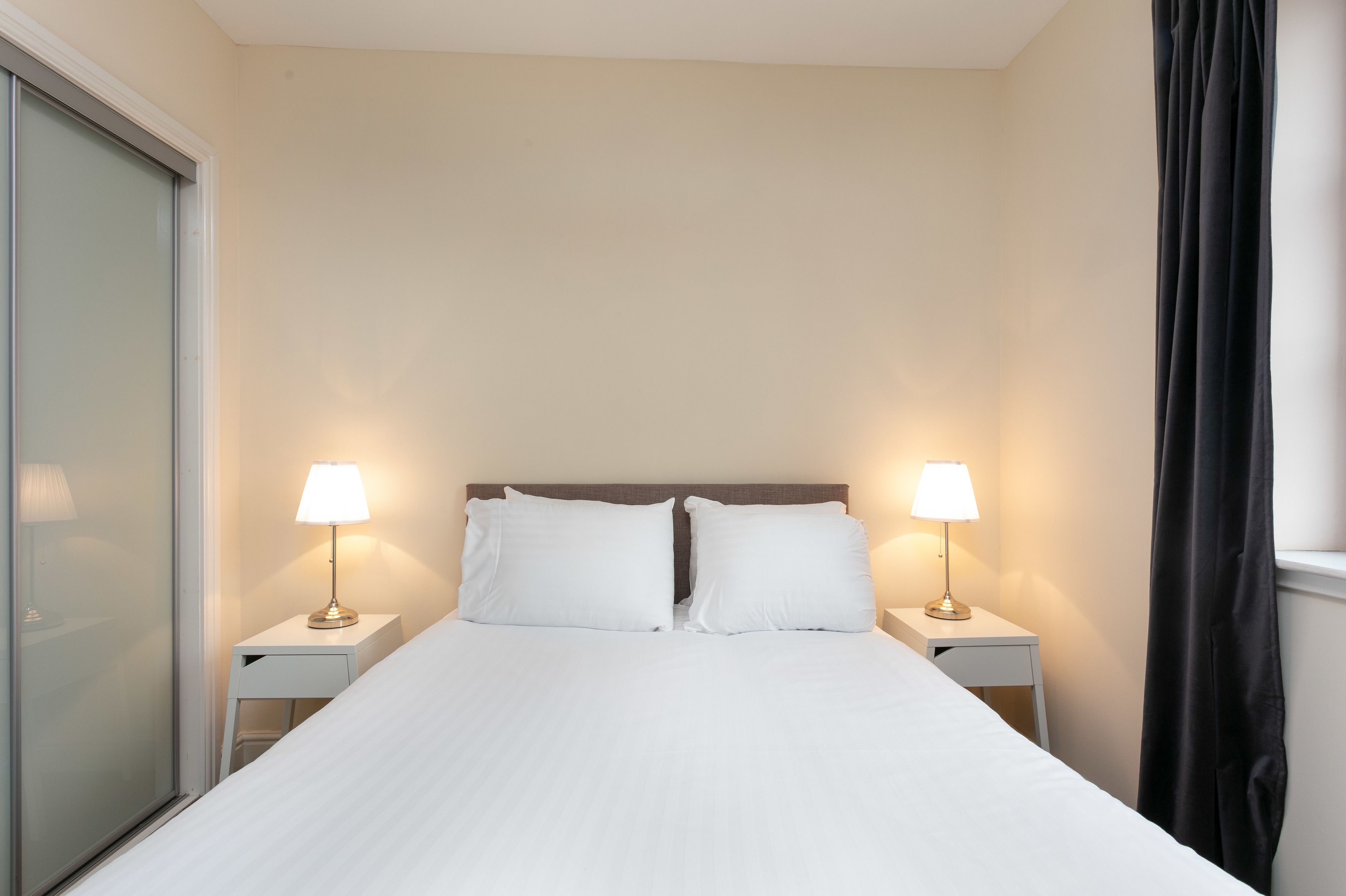 Cama bem arrumada com lençóis brancos, duas luminárias de cabeceira e uma janela com cortinas pretas.