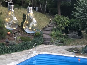 A swimming pool with a stone border, surrounded by a garden with steps leading up to a house. - Haus Am Wald (Eltmann)