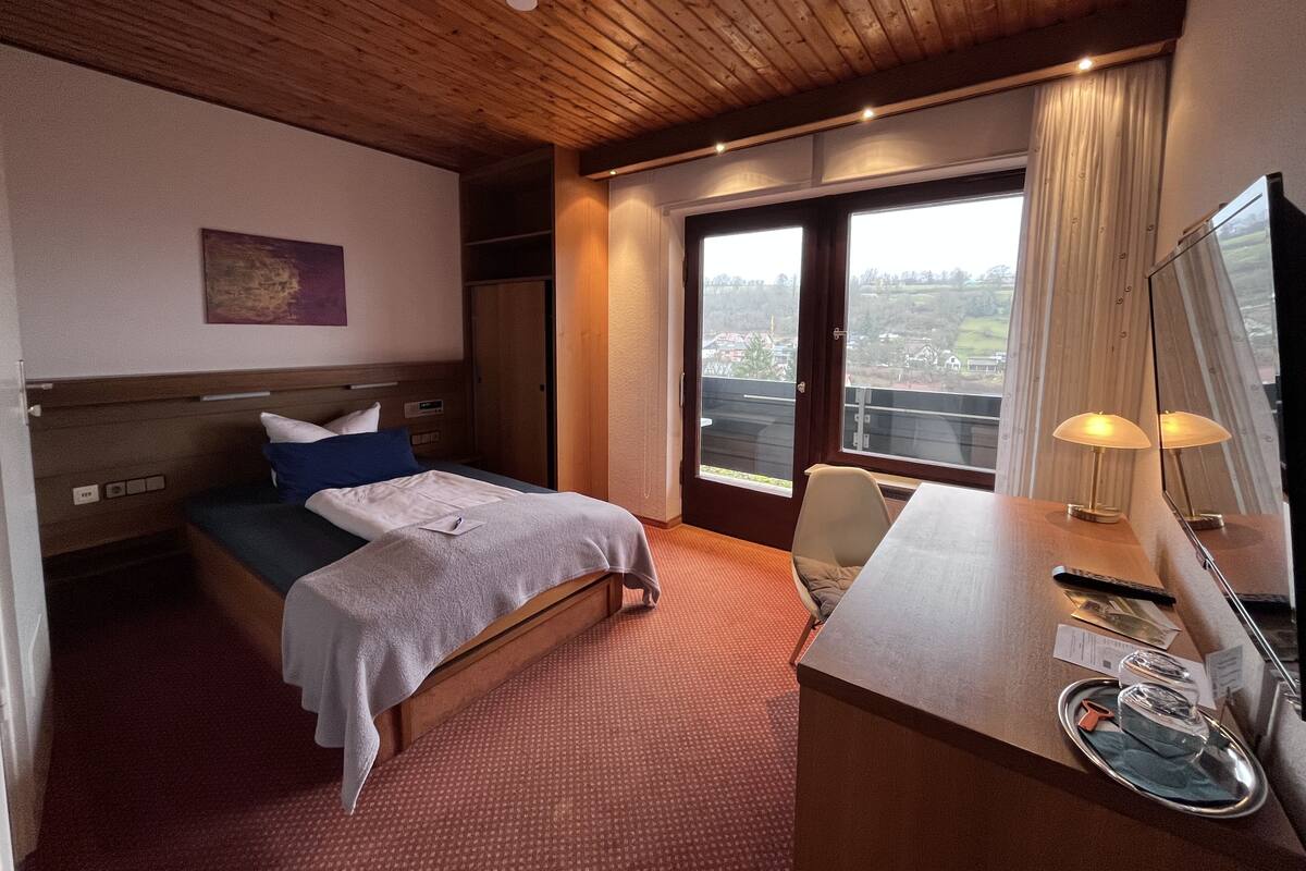 A hotel room with a bed, a desk with a lamp, a large window with a view, and a mirror.