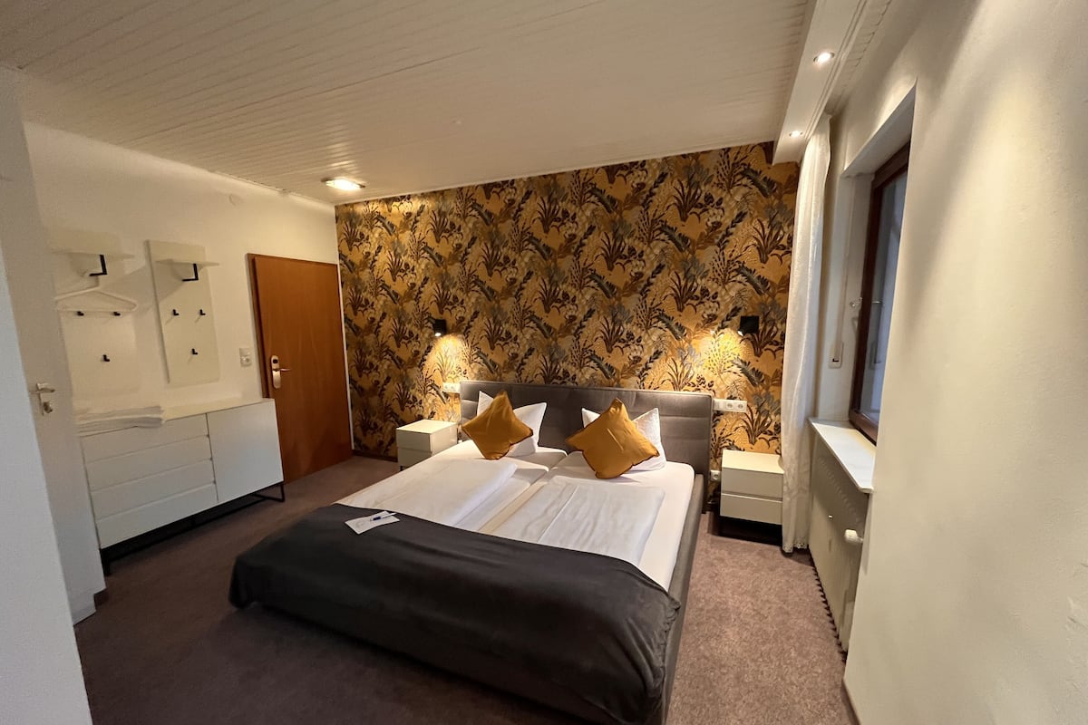 A hotel room with a large bed, two bedside tables with lamps, a wardrobe, and a patterned wallpaper.