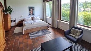 Un dormitorio con cama, una silla, una mesa, un ventanal con cortinas, una planta y un cuadro en la pared.