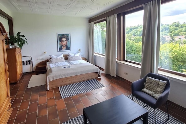 A bedroom with a bed, a chair, a table, a window with curtains, a plant, and a painting on the wall. - Haus Am Wald (Eltmann)