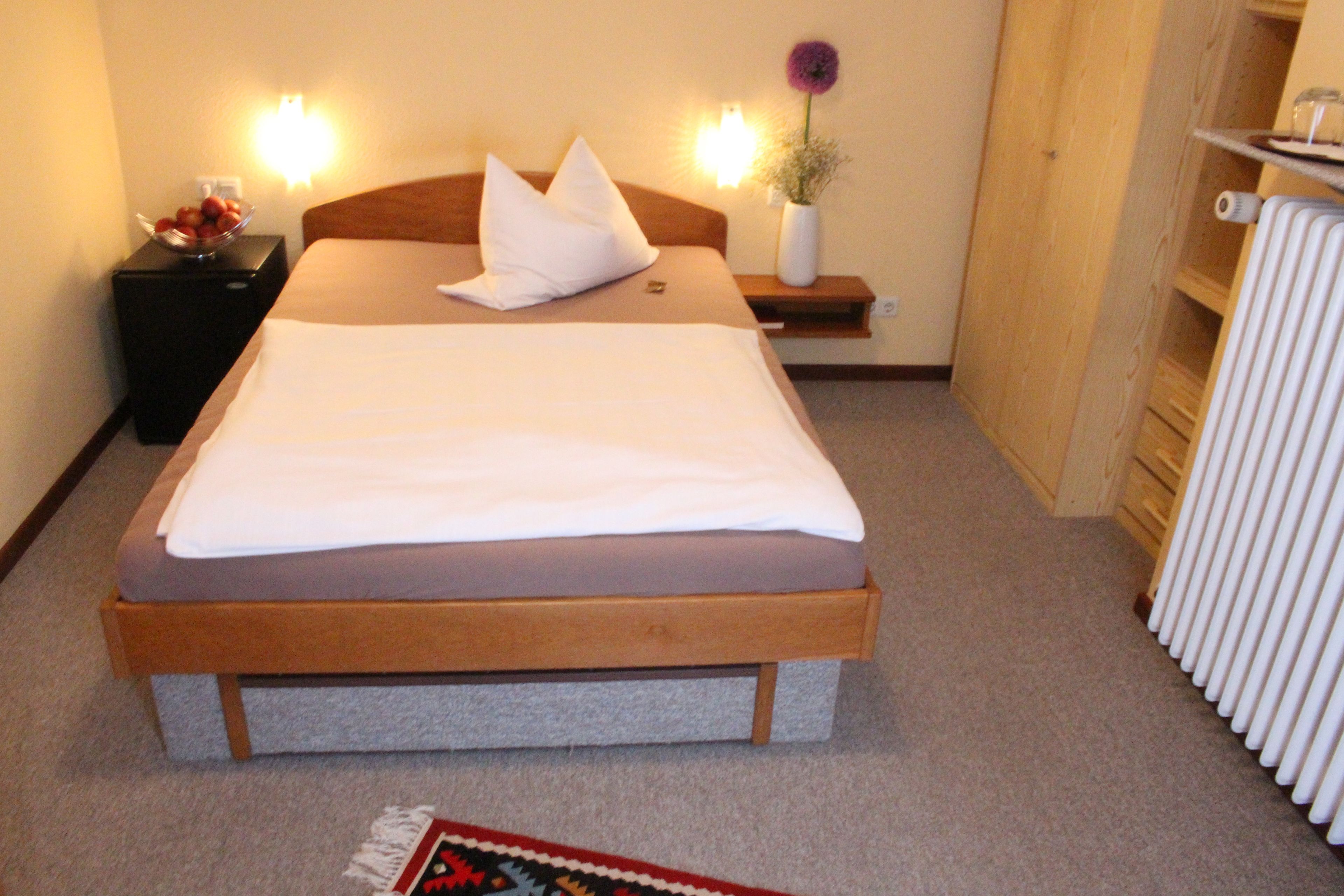 A neatly made bed with a wooden frame, a bedside table with a bowl of fruit, a vase with a flower, and a wall-mounted light.