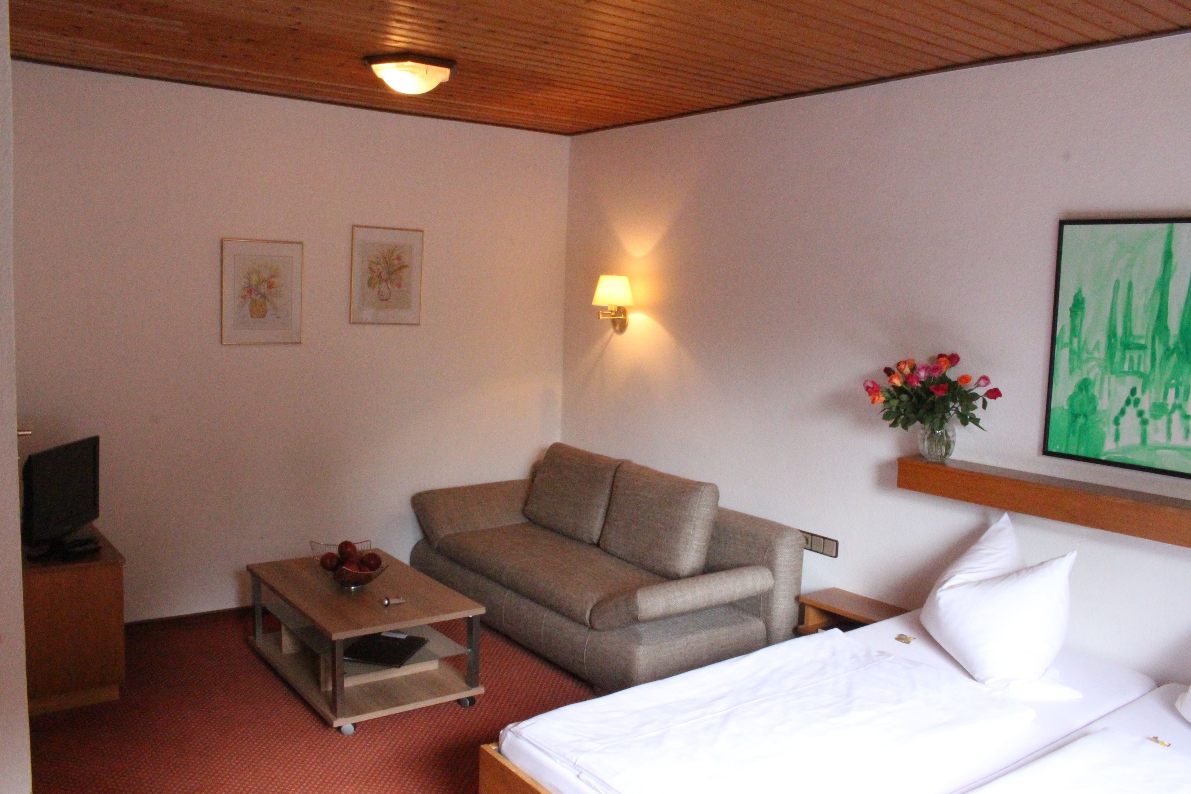 A hotel room with a bed, a sofa, a small table with a bowl, a TV on a wooden cabinet, and a wall-mounted lamp.