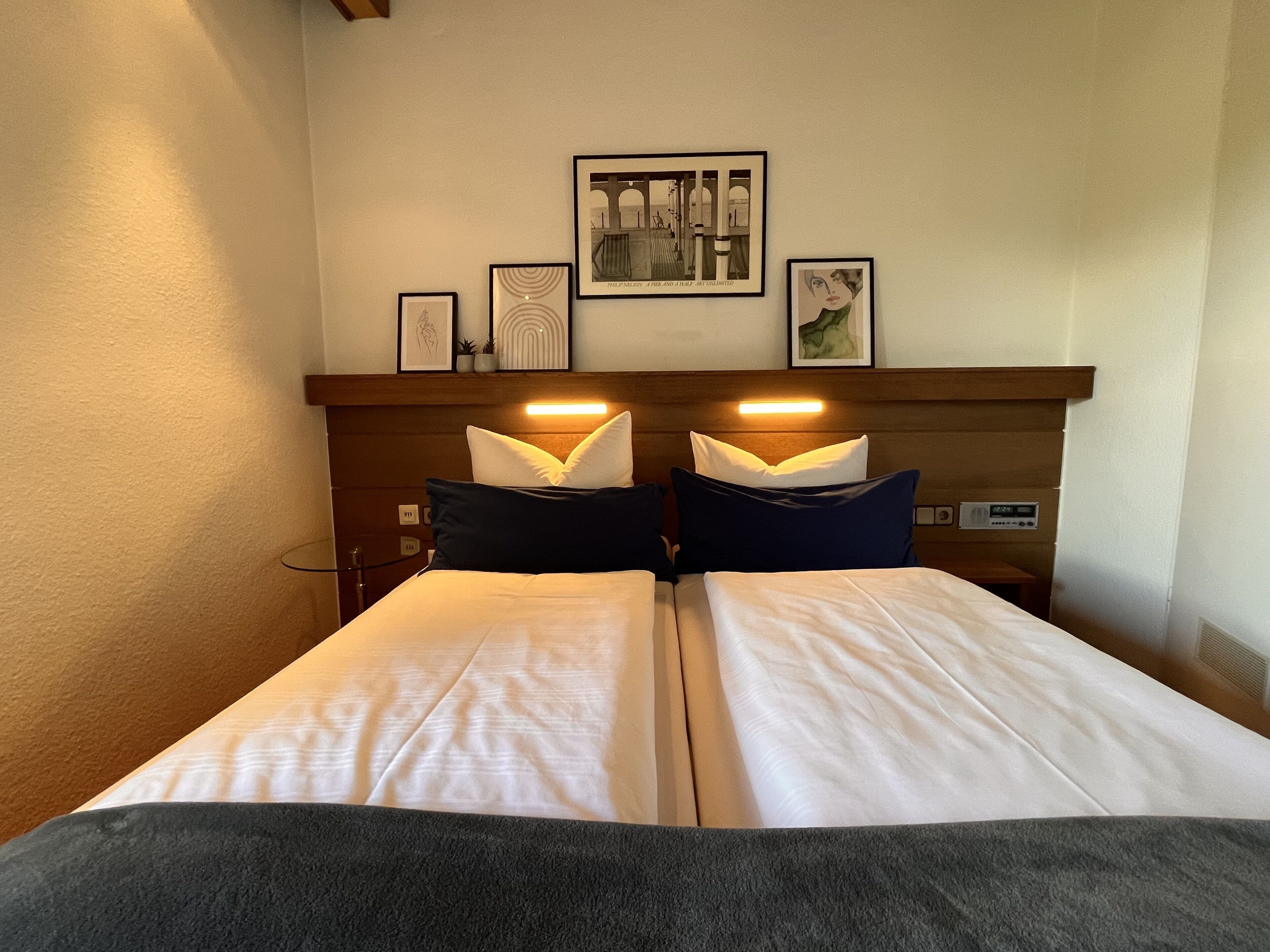 A hotel room with a bed, two bedside tables, a nightstand, a wall-mounted shelf with framed pictures, and a wall clock.