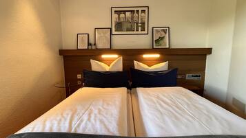 A hotel room with a bed, two bedside tables, a nightstand, a wall-mounted shelf with framed pictures, and a wall clock.