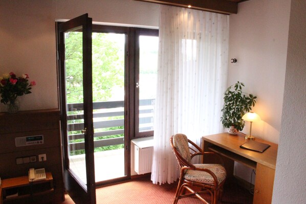Une pièce avec un bureau en bois, une chaise, une plante en pot, une lampe et une vue sur l’extérieur, visible à travers une porte-fenêtre coulissante.