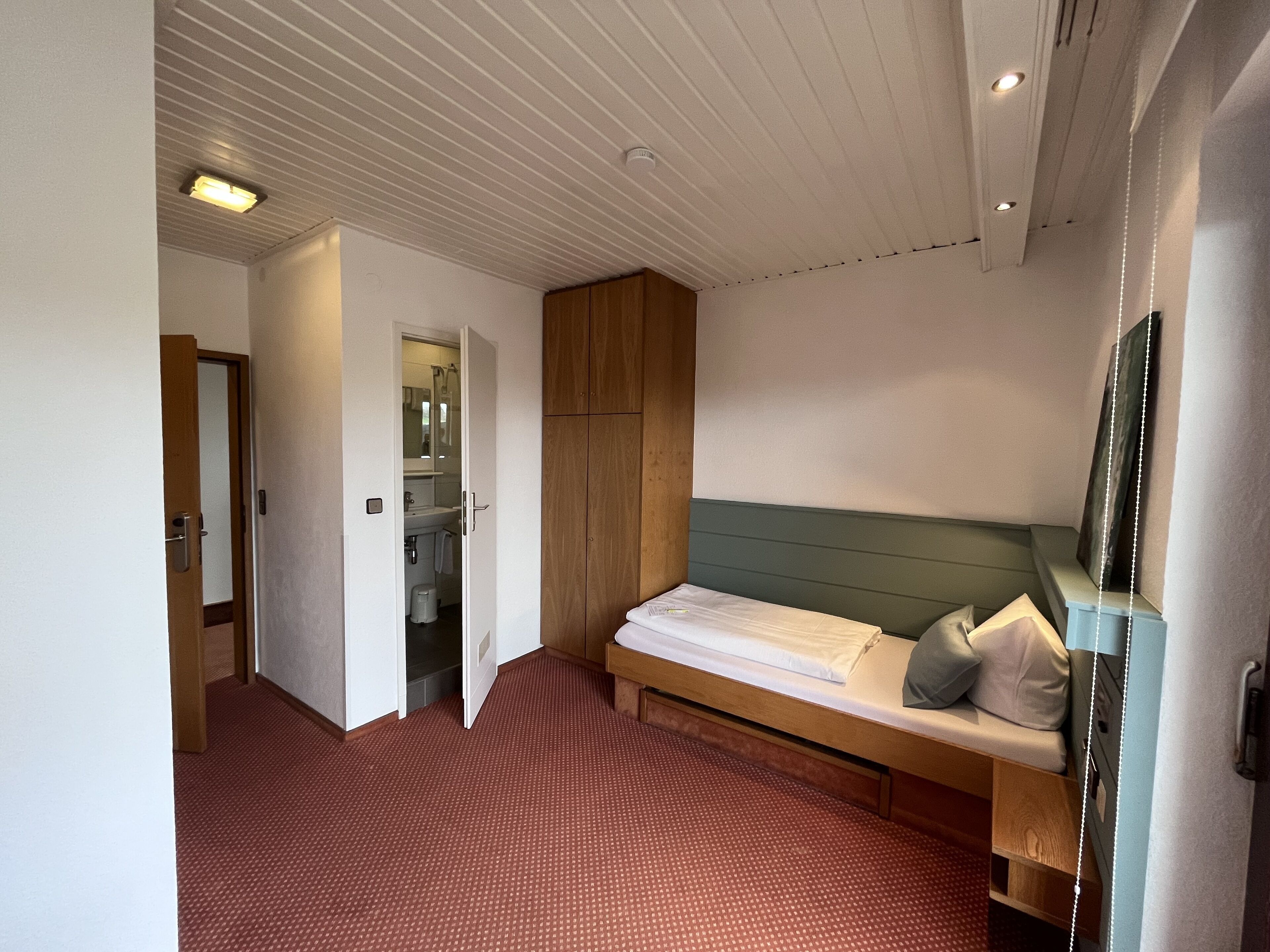 Camera da letto con un letto singolo, un armadio in legno e un bagno visibile attraverso una porta aperta.