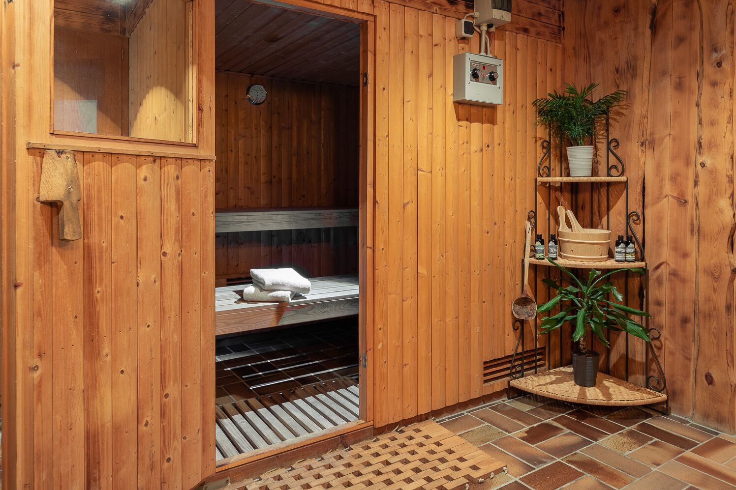 A wooden sauna room with a bench, towels, a clock, a control panel, and shelves with plants and bottles.