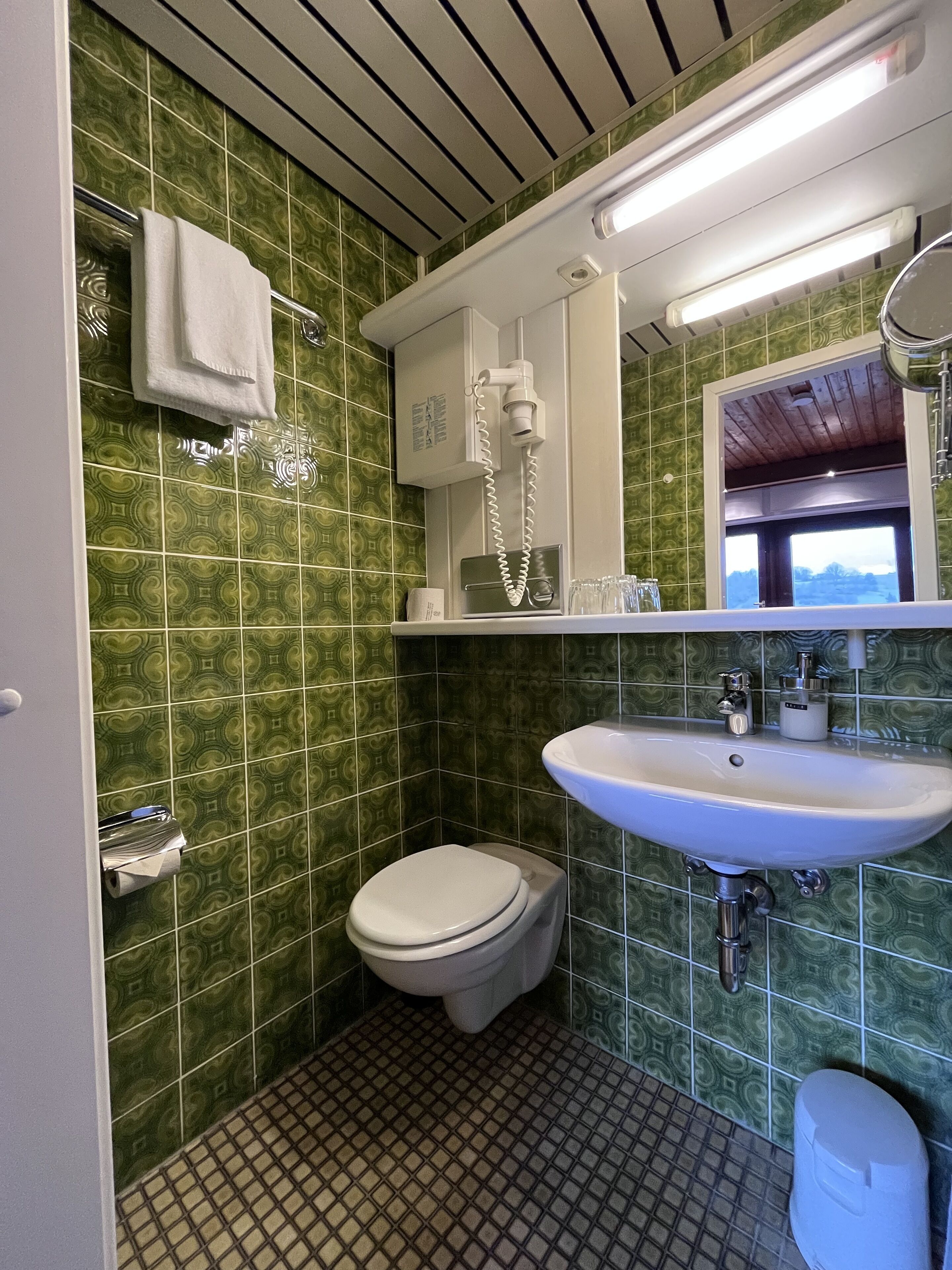 Un bagno con piastrelle verdi a motivi, un lavandino bianco, un water e un asciugamano appeso al muro.
