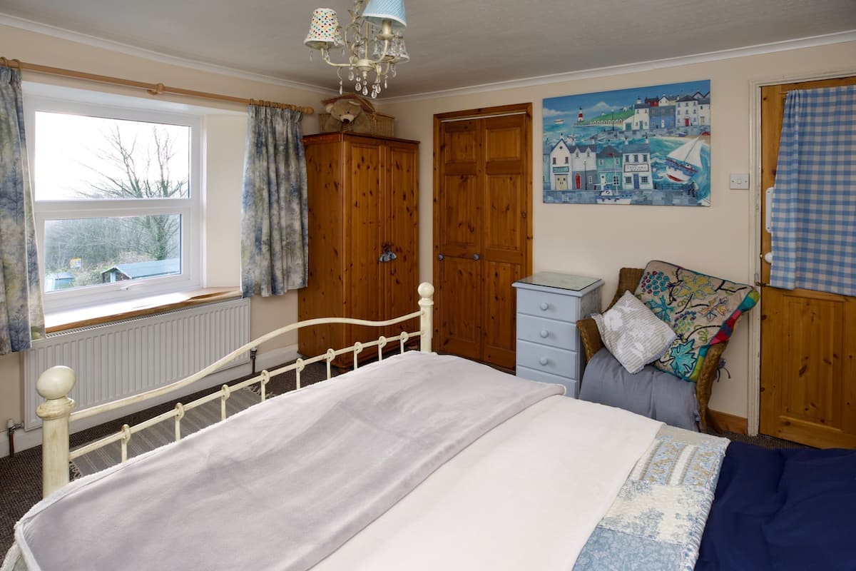 A bedroom with a bed, a dresser, a nightstand, a window with curtains, a wooden wardrobe, and a painting on the wall.