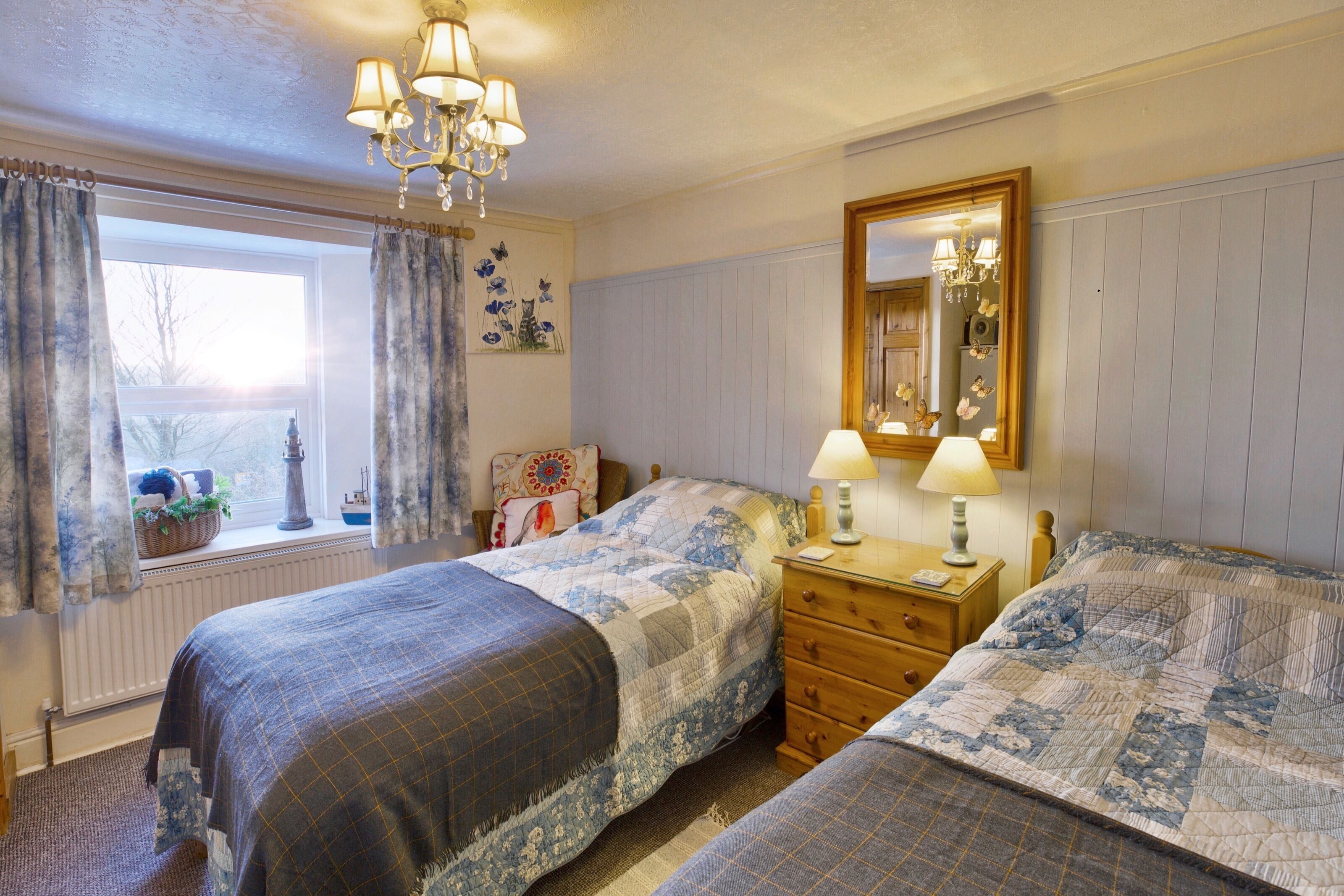 A bedroom with two beds, a dresser, a nightstand, a window with curtains, a chandelier, and a mirror.